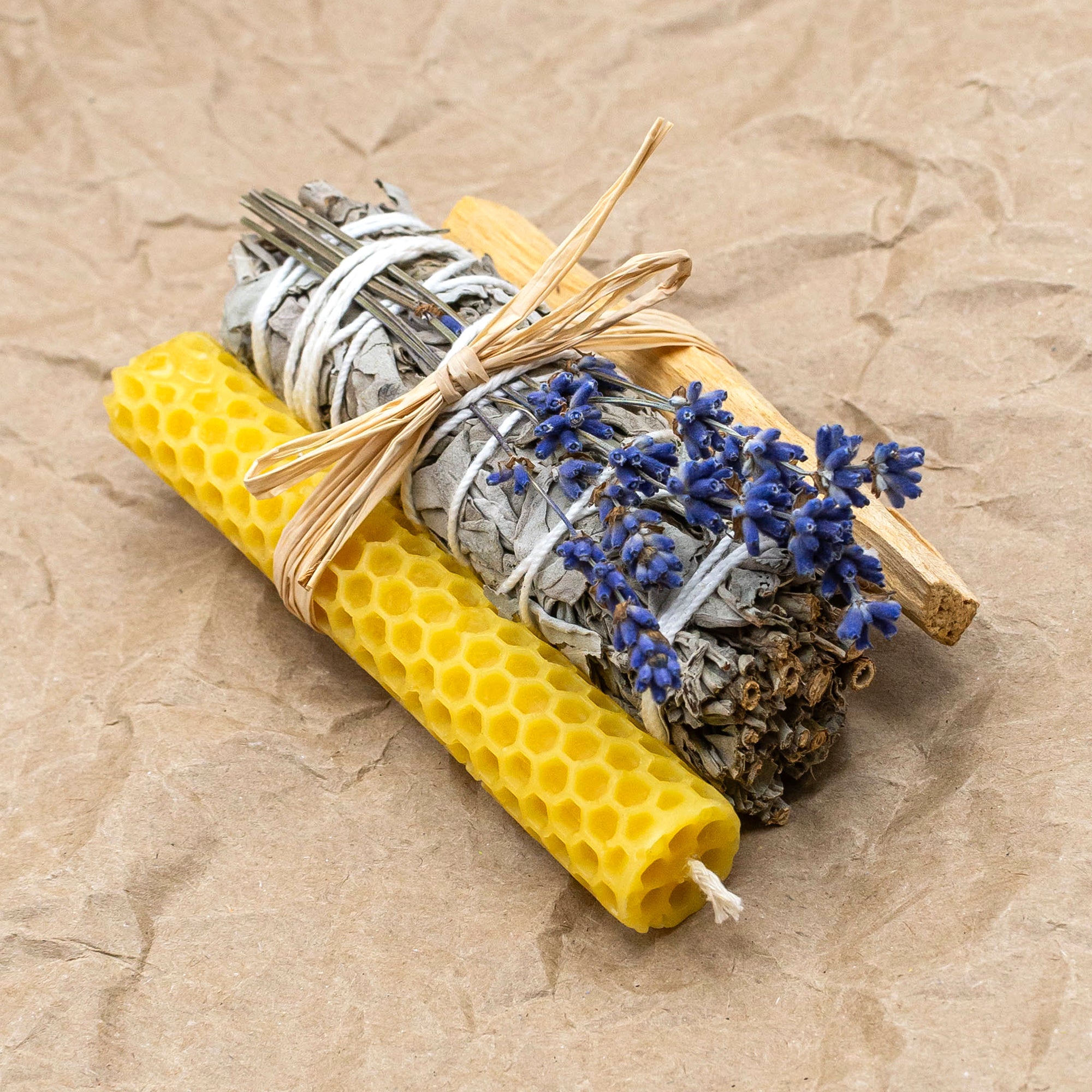 white sage bundle, with a honeycomb textured beeswax candle and a stick of palo santo, and a bunch of purply blue lavender. All tied together with a piece of raffia. The bundle sits on some crinkled brown paper.